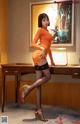 A woman in an orange dress and black stockings posing in front of a desk.