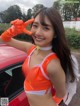 A woman in an orange and white outfit posing next to a red car.