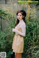 A woman standing in front of tall grass wearing a beige dress.